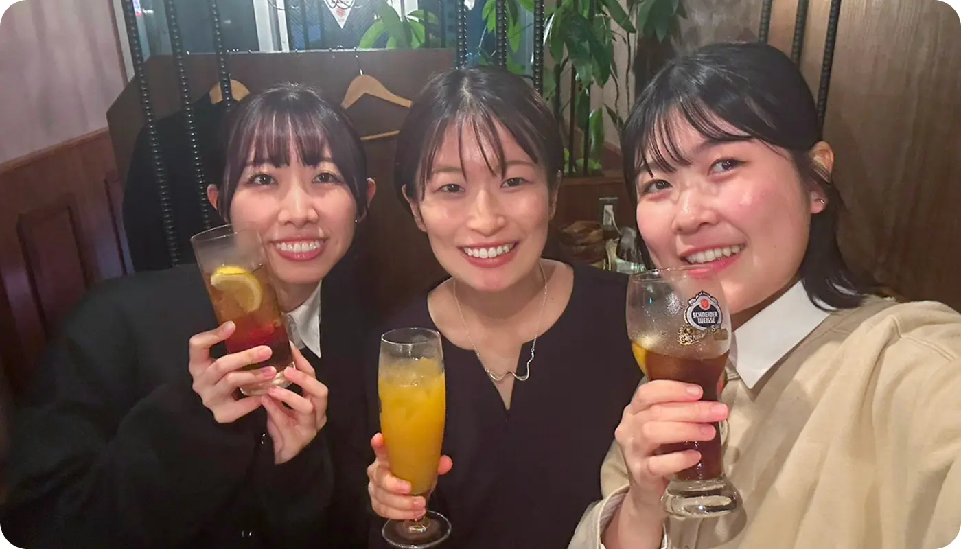 親睦会（食事会）でドリンクを手に笑顔で並ぶ、仲の良い女性スタッフ3名のオフショット。